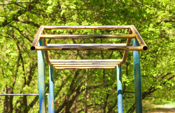 Outdoor monkey bars in a sunlit park with green spring trees in the background, showing weathered metal playground equipment for exercise and recreation
