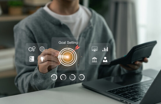 Businessman using virtual interface with digital icons and graphic charts focusing on goal setting concept in modern office while holding calculator and working on laptop computer