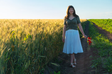 Obraz premium Young woman walking in wheat field holding red flowers at sunset