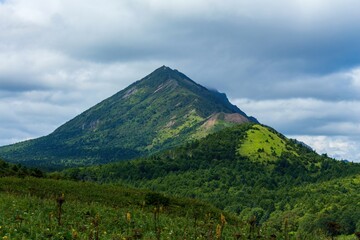 Fototapeta premium A majestic green mountain with a distinct pyramidal peak rises above a dense forest, its slopes covered in vibrant vegetation, with a dramatic sky of heavy grey clouds overhead