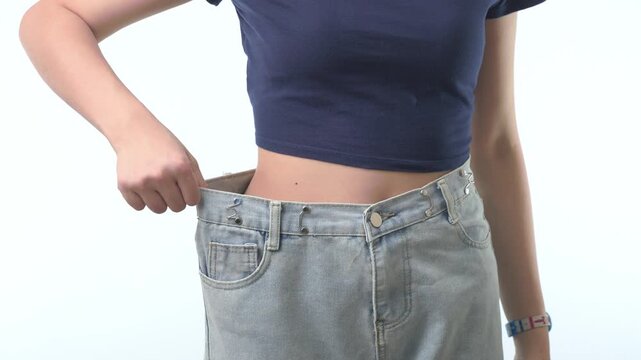 Proud slim woman showcases successful healthy weight loss journey, pulling loose oversized blue jean to reveal fit belly against white background