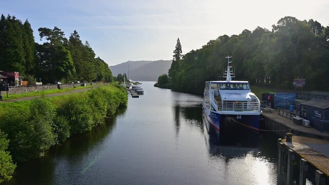 Ein ruhiger Fluss mit Booten, umgeben von Wald und sanften H&uuml;geln unter einem klaren Himmel, Kaledonischer Kanal, Fort Augustus, Loch Ness, Schottland, Vereinigtes K&ouml;nigreich, Gro&szlig;britannien