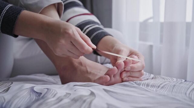 Woman sitting on a bed filing her toenails with an emery board, performing an at‑home pedicure and foot grooming as part of a self‑care routine in a cozy, natural‑light bedroom setting.