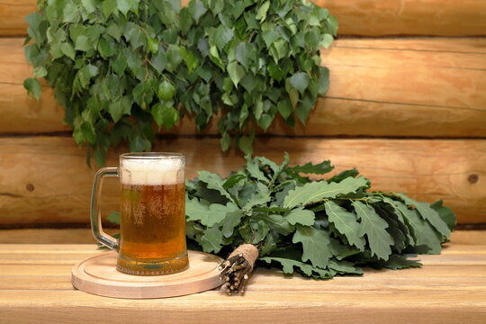 A mug of light beer on a round wooden tray next to a fresh oak broom stands on a plank bench against the background of a log wall with a couple of fresh birch brooms hang on it.