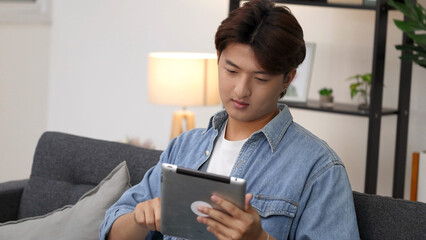 Young Asian Man in Denim Shirt Using Tablet on Living Room Sofa © blueringmedia
