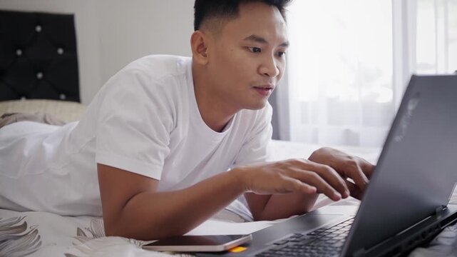 Young Asian Man Working or Studying Remotely on Laptop in Casual Bedroom Workspace Concentrating on Screen