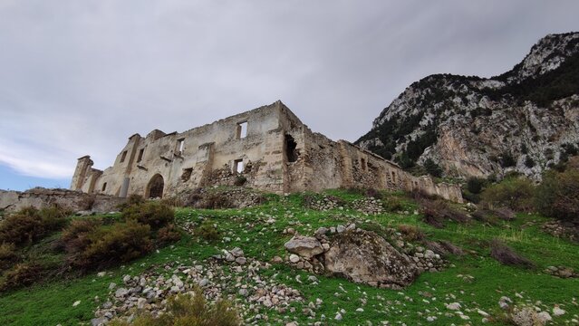 A low-angle exterior view of the historic and abandoned Sina Monastery, also known as the Vasilya Agia Paraskevi Monastery, set against a mountainous landscape under a cloudy sky.