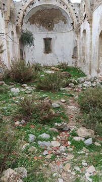 View inside the ruined interior of the Sina Monastery (Vasilya Agia Paraskevi Monastery), showing crumbling stone walls, a vaulted arch, and vegetation growing on the floor.