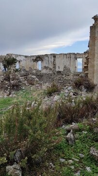 View of the crumbling stone ruins and remaining walls of the historic Sina Monastery (Vasilya Agia Paraskevi Monastery), surrounded by wild vegetation.