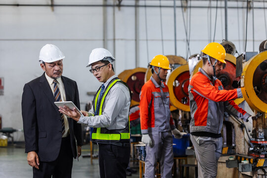 Factory manager and manufacturing engineer reviewing production performance using a digital tablet while technicians operate industrial press machines on the production line.