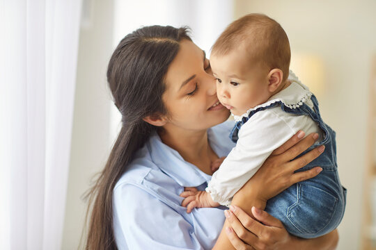 A smiling mother holds her baby by the window, sharing a gentle kiss and affectionate bonding in a bright home portrait that conveys maternal joy. Mother kisses her toddler with gentle care