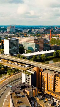 Vertical video. Ghent, Belgium. Esco (Scheldt) river embankment, aerial panoramic sweep over the waterway, tree-lined banks, and dense urban rooftops under a cloudy summer sky. Drone footage, Departu