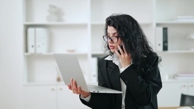 Professional woman in glasses expertly juggles phone call, laptop work. Displays diligence, commitment handling office tasks with focused energy, conveying serious engagement.