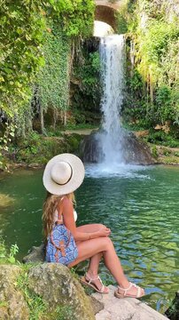 Woman sitting by tropical waterfall looking at cascade in forest, Tobera. Burgos, Castilla y Leon. Spain