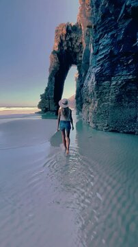 Woman walking on sandy beach under massive natural rock arch, Playa las catedrales Catedrais beach in Galicia, Spain