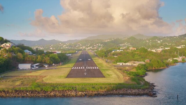 Scenic view of airport runway extending toward lush green mountains in Saint Maarten. Tropical landscape with residential hillside homes, calm blue ocean water, and dramatic clouds at golden hour.