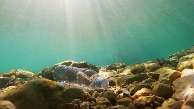 A plastic bottle rests on the ocean floor among rocks. Sunlight filters through the water. Marine plastic pollution impacts ecosystems and wildlife. Awareness is needed to protect our oceans.