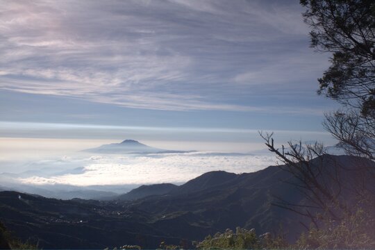 Panoramic mountain landscape with sea of clouds, distant volcanic peak, and soft morning light over silhouetted hills in a tranquil, hazy atmosphere.