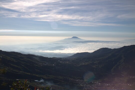 Panoramic mountain landscape with distant peak above sea of clouds. Atmospheric morning light over misty valley and volcanic range under blue sky.