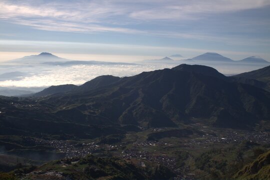 Panoramic mountain landscape at dawn with sea of clouds, distant volcanic peaks, and misty valley village under soft morning light.