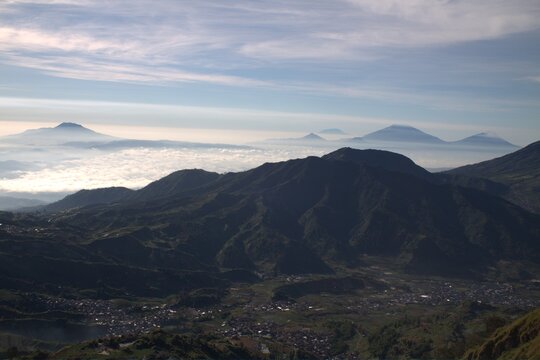 Panoramic mountain valley view with village, distant volcanic peaks above morning mist, soft daylight, serene atmosphere.