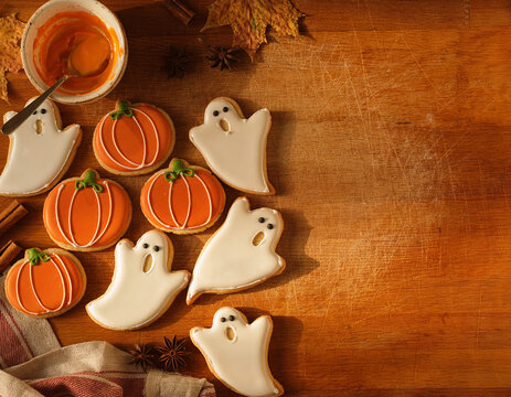 Halloween pumpkin and ghost shaped iced sugar cookies on wooden background with autumn decorations and baking ingredients, festive seasonal holiday treat concept