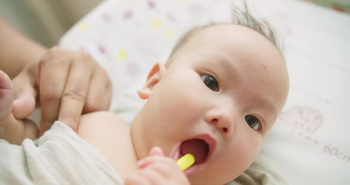 A curious Asian infant learns about oral hygiene for the first time, chewing on a baby toothbrush while a parent provides gentle assistance and care in a home setting.