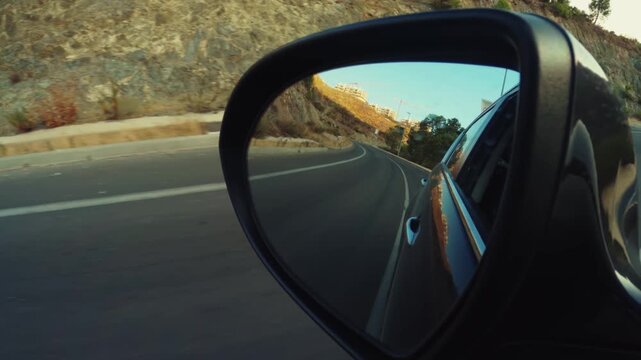 View from car side mirror during dynamic drive on mountain road with beautiful reflection of sky and landscape.