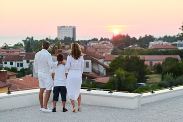 Obraz premium Family On Rooftop Terrace Watching Sunset Over Coastal Town And Sea View