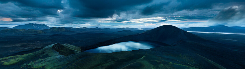 Fototapeta premium Panoramic view of Ljótipollur crater lake in Iceland's Highlands, surrounded by dark volcanic slopes beneath dramatic storm clouds. A remote and breathtaking volcanic landscape.