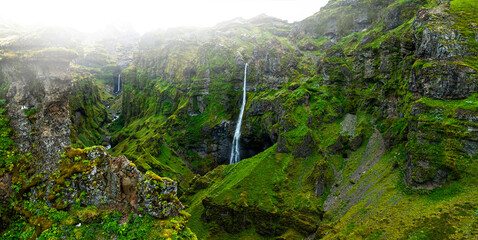 Panoramic view of the breathtaking Múlagljúfur Canyon in southeast Iceland, featuring dramatic moss-covered cliffs, a cascading waterfall, and a winding river beneath soft mountain light.  © Jag_cz