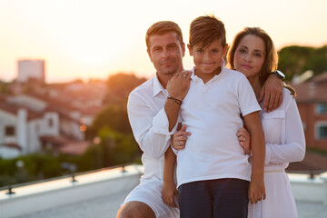 Obraz premium Happy Family on Rooftop Balcony at Sunset Embracing Son With Sea View and Warm Togetherness