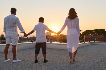 Obraz premium Family Holding Hands on Rooftop Terrace at Sunset With Sea View and Coastal Town Background