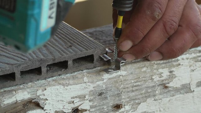 Professional deck installation involves a worker securing composite planks with metal clips using an electric power drill. Extreme close-up shot.
