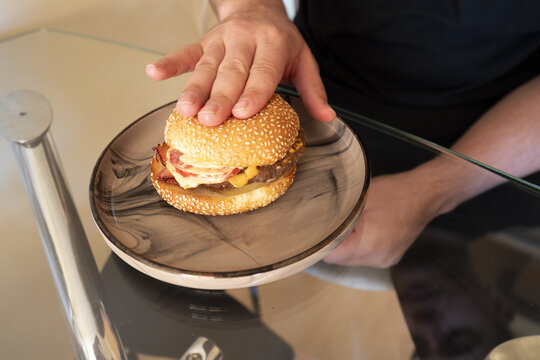 Hand pressing a delicious hamburger on a stylish plate, showcasing layers of meat, cheese, and toppings ready to be enjoyed.