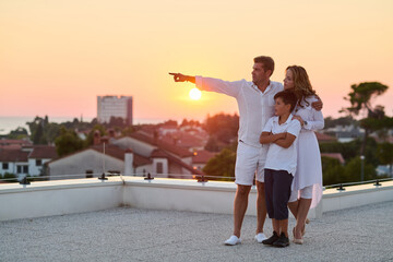 Obraz premium Family on Rooftop Balcony at Sunset With Sea View, Parents Pointing Toward Horizon