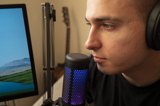 A focused young man wearing headphones speaks into a microphone while recording in a home studio with a computer and guitar in the background.