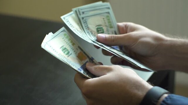 Hands hold a stack of United States hundred dollar bills while counting currency to complete a financial transaction in a professional business setting with a neutral background.