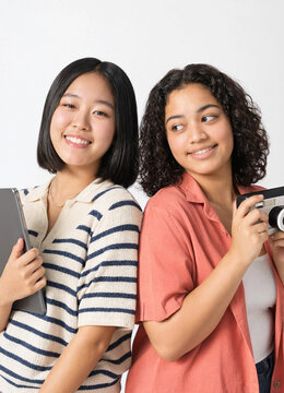 Two young diverse female students standing back to back with tablet and camera on white background