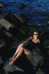 Fototapeta premium Woman relaxing on rocks by the water during daylight, wearing black swimwear and sunglasses while enjoying the outdoor setting
