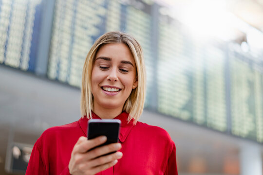 Smiling young businesswoman using cell phone at arrival departure board at the airport