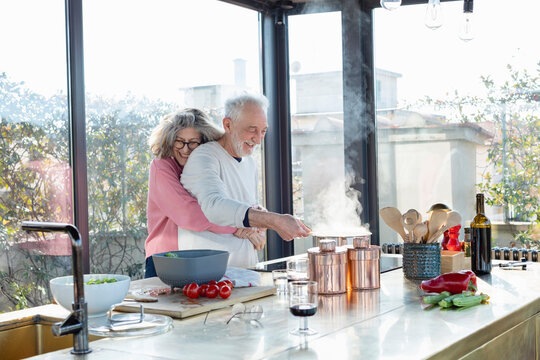 Smiling senior woman embracing man from behind in kitchen at home