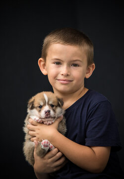 Close-up vertical portrait of a cute young boy cuddling a baby Pembroke Welsh Corgi puppy against a dark background.