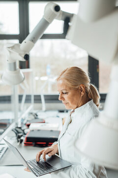 Scientist using laptop by robotic arms in laboratory