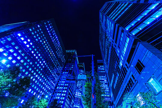 Dynamic low-angle looking up perspective of futuristic corporate skyscrapers lining a dark night sky canyon in Tokyo.