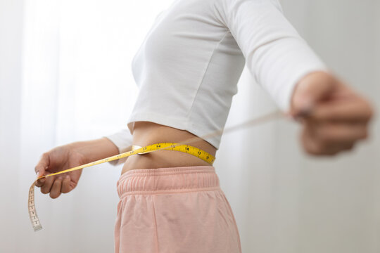 Young woman measuring slim waist with tape measure. Focused female checking her body size and progress for weight loss, part of diet and healthy lifestyle indoors