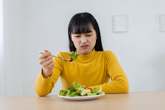 Unhappy young Asian woman looking at food with disgusted expression. Reluctant girl on diet eating healthy salad for new lifestyle, feeling bored and picky