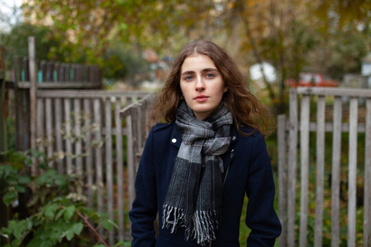 Cheerful woman in a coat and scarf stands outdoors by a wooden fence