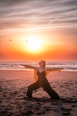 Woman in the warrior two yoga pose with extended arms on the beach while birds drift across the sunset sky a serene scene of strength focus and mindful movement. © Oksana