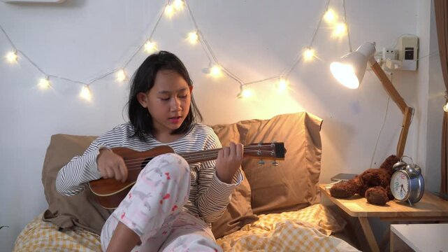 Young girl sits comfortably on her bed practicing the ukulele after her online classes in bedroom illuminated by warm fairy lights
